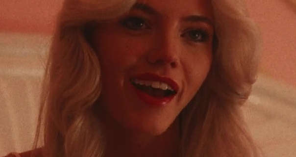 Hayes Warner sits on a large bed in front of a white and pastel pink headboard with her blonde hair loose and in soft waves. She wears a light foundation with black mascara and a shiny red lipstick.