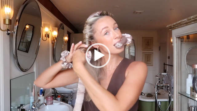 Fashion Mumblr stands in her white and dark green bathroom at her home. She wraps her hair around a taupe sausage to create heatless curls. Josie wears a brown top with foundation, mascara, pink lipstick, small earrings and a think necklace.