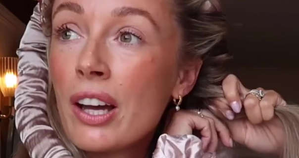 Josie Irons looks up and to the side as she works through a hair routine. Josie stands near an glass lamp in her bathroom at home. She winds her hair around a heatless curler sausage. Josie wears a full foundation with mascara, pink lipstick, gold rings and a pair of pearl drop earrings.