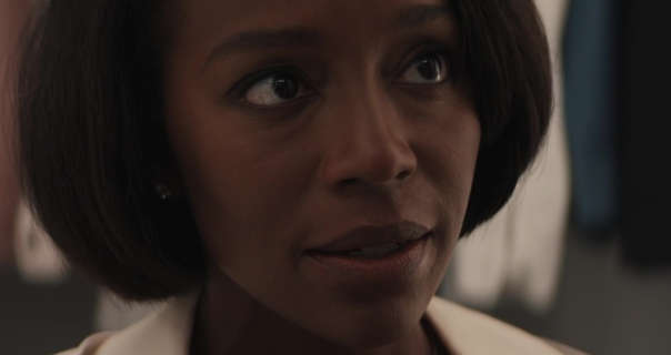 Aja Naomi King stands in a large walk in dressing room. She has her short, thick black hair loose and straightened. Aja wears foundation with black mascara, dark mauve lipstick, small earrings and a cream top.