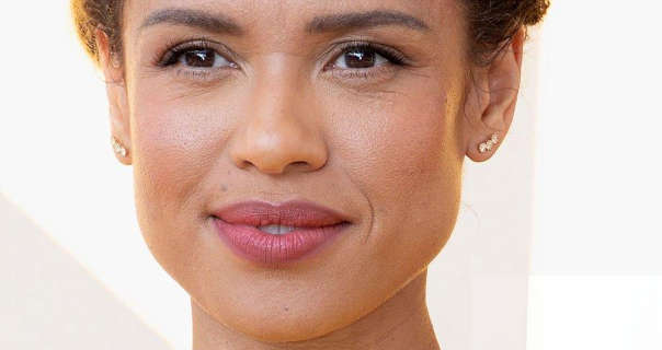 Gugu Mbatha-Raw is seen in front of a pale grey and white event wall. She has her curly hair tied back. Gugu wears a light coverage foundation with blush, mascara, dark pink lipstick and a few diamond stud earrings.