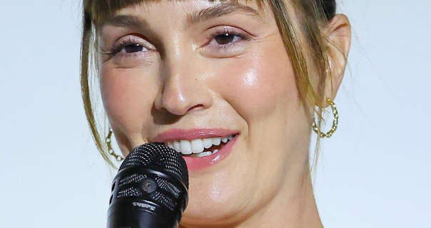 Leighton Meester is seen in front of a dark white wall. She holds a large black microphone to her face as she appears on stage. Leighton has her long hair tied up with strands pulled down to frame her face. She wears a shiny foundation with mascara, medium size gold hoop earrings, blush and a light pink lipstick.