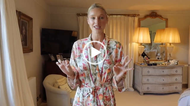 Leonora Smee pinches her fingers and thumbs together on both hands to accentuate a talking point. She stands in her filming bedroom at her countryside home. Leonora wears her long fruit print dressing gown with a full foundation, blush, mascara and a dark pink lipstick. She has her long blonde hair tied back in a bun.