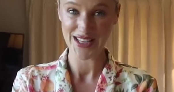 Leonora Smee appears in her bedroom at home in front of her closed cream blinds. Leonora has her long blonde hair tied back in a tight bun. She wears a long white, green, orange and pink dressing gown. Leo appears in a dewy foundation, mascara, black eyeliner and a dark pink lipstick.