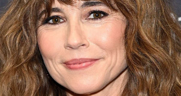 Linda Cardellini is seen in front of a light grey event wall that has white text and logos across it. Her brown hair is loose and in a natural frizz. Linda wears black eye liner with mascara, pink lipstick, matt foundation and a pink satin lipstick.