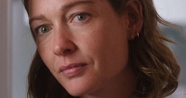 Cristiana Capotondi is seen with her hair loose, in natural waves and cut to around shoulder hair. She leans against a white kitchen doorframe in a small apartment during the day. Cristiana wears light makeup with a pink lipstick and small gold circle earrings.