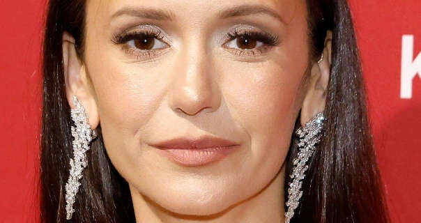 Nina Dobrev stands in front of a dark pastel red wall that has white display text across it. She has her long black hair straightened and loose. Nina wears a reflecting foundation with mascara, dark pink lipstick and a pair of hanging diamond encrusted earrings.