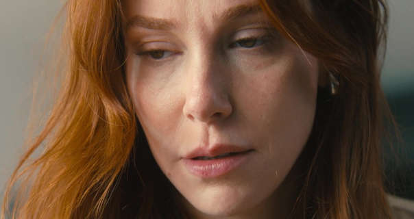 Elçin Sangu sits in an open plan kitchen area during the day. She has her long red hair loose and in natural waves. Elçin wears a light coverage foundation with blush, pale pink lipstick and a pair of thin silver earrings.