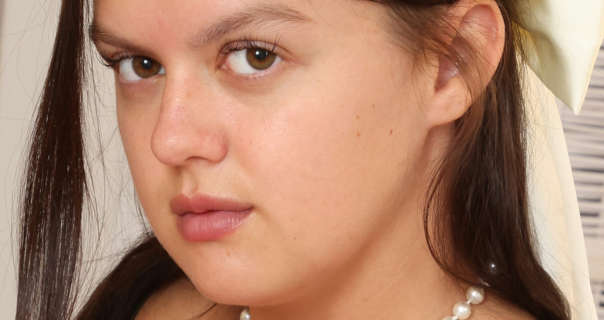 Kelsey is seen with her long dark brown hair loose and pinned back with a large white bow. She kneels on a bed in front of a white closet and a white wicker changing screen. Kelsey wears a light coverage foundation with curled lashes, dark pink lipstick and a pearl necklace.