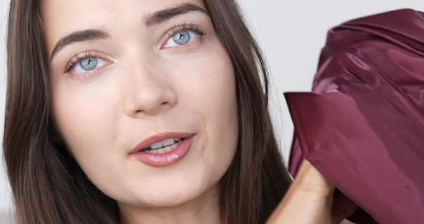 Roxxsaurus is seen with her long brown hair loose and naturally straight. She holds up a maroon Shein dress as she talks to the camera. Roxi wears a matt foundation with a little pink eye shadow, blush and a dark pink lipstick. She sits in front of a dark white wall.