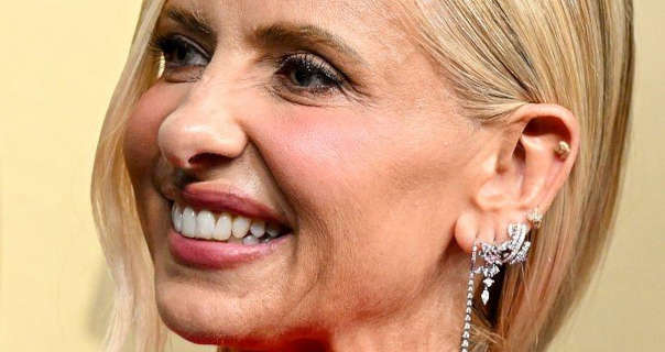 Sarah Michelle Gellar stands in front of a light gold gradient wall. She has her long blonde hair tied back in a low ponytail. Sarah wears a natural foundation with pink blush, mascara, dark pink lipstick, gemstone covered dangling earrings and gold ear studs.
