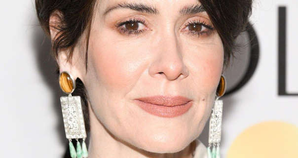 Sarah Paulson stands in front of a dark white event well that has thin black text and light gold circle on it. She has her black hair cut short and tied back. Sarah wears a full foundation with blush, smoky red eye shadow, black mascara, dark pink lipstick and a pair of tiger eye gemstone tassel earrings.