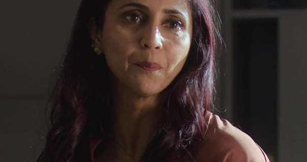 Gautami Kapoor stands in a fairly dark living room with sunlight hitting her side. She is seen with her long black hair loose, styled naturally and with red tints. Gautami wears a light foundation with mascara, several earrings, dark red matt lipstick and a pastel brown shirt.