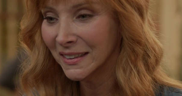 Lisa Kudrow is seen with her long light orange hair loose and in waves. She stands outdoors in a film lot as the sun sets. Lisa wears a soft foundation with light pink blush, mascara and a baby pink lipstick.