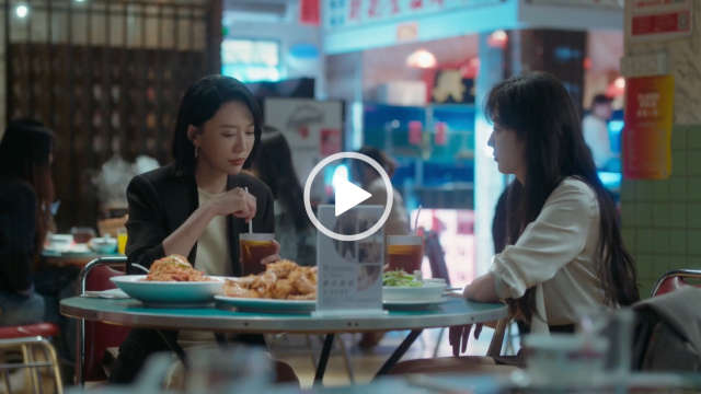 Sun Qian sits at a circular table at the indoor food market with a friend. She has her long black hair loose and styled naturally. Sun Qian wears a light gold skirt with a short skirt. She sits with