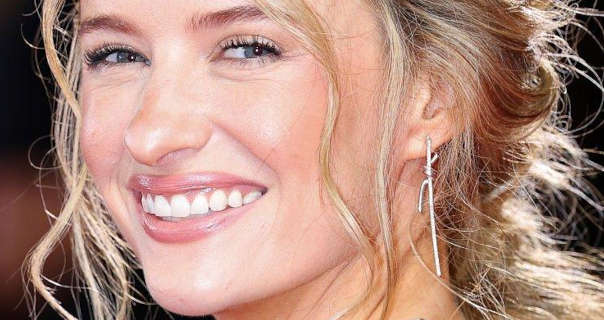 Victoria Magrath smiles as she appears at a nighttime red carpet event. She has her long blonde hair tied back in loose plaits. Victoria wears a dewy foundation with mascara, silver wire earrings, blush and a pink sheen lipstick.
