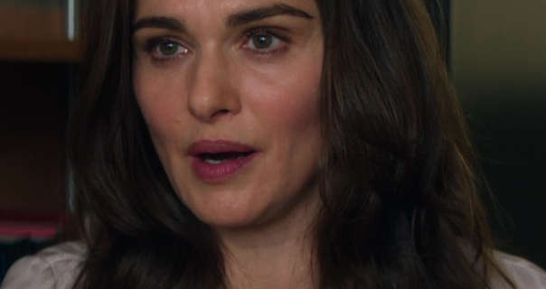 Rachel Weisz looks to one side as she talks. She sits on the floor of a small office with black frame wood shelves and a green wall behind. Rachel wears a dewy foundation with blush, mascara, dark pink lipstick and a long sleeve cream top. She has her thick dark brown hair loose.