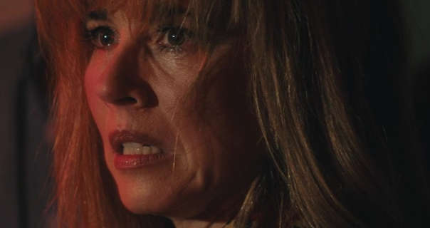 Linda Cardellini appears in a dark room in front of a deep brown wooden door. She has her thick, long hair loose and with bangs. Linda wears a light coverage foundation with black mascara and a dark pink lipstick. She looks forwards with a fierce and slightly unnerved expression.