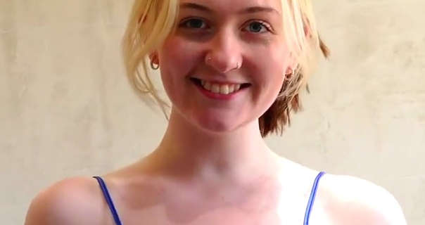 Em Yang smiles and looks into the camera as she stands in a cream tiled bathroom. Em has her long blonde hair tied back with some strands framing her face. She wears a matt foundation with natural blush, small gold hoop earrings, mascara and a dark pink lipstick.